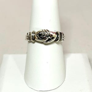 May include: A silver-toned ring featuring a design of two hands clasped together at the centre. The band has a polished metallic finish with subtle dark oxidation in the recessed areas to highlight the intricate details of the fingers and cuffs.