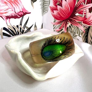 以下が含まれることがあります： 半透明のベージュ色の樹脂製ワイドバングルブレスレット。鮮やかな緑、青、黒の色合いで描かれた、手描きのクジャクの羽のデザインが特徴です。滑らかで光沢のある仕上げが施されており、湾曲した円筒形の形状をしています。