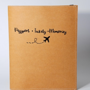 Könnte beinhalten: Ein rechteckiger Reiseunterlagenhalter in Beige mit dem schwarzen Text Passport • Tickets • Memories und einer schwarzen Grafik eines Flugzeugs mit einer gestrichelten Flugbahnlinie. Die Hülle hat sichtbare Nähte an der rechten und oberen Kante.