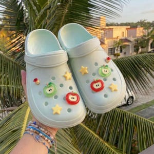 May include: A pair of light mint green foam clogs featuring a series of circular ventilation holes. The clogs are decorated with various charms, including red and green apple shapes and small yellow stars attached to the upper surface of the shoes.