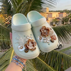 May include: A pair of light mint green slip-on clogs featuring decorative embellishments on the top. The adornments include brown resin flowers, clear crystal-like floral accents, silver-toned metallic spikes, and smooth brown circular beads.