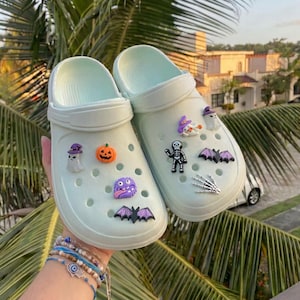 May include: A pair of light mint green slip-on clogs featuring various Halloween-themed charms. The charms include a ghost with a purple hat, an orange pumpkin, a purple monster, a bat, a skeleton, a white ghost, and a skeletal hand.