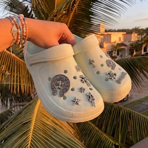 May include: A pair of off-white foam clogs decorated with various silver-toned metal charms, including cross, star, and fleur-de-lis shapes, along with a circular emblem featuring the text Chrome Hearts, all attached to the ventilation holes.