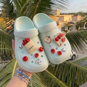 May include: A pair of light mint green slip-on clogs featuring various decorative charms. The charms include a paper bag labeled TOMATO, red tomato shapes, a wooden cutting board with sliced tomatoes, and small silver-toned star and heart accents.