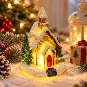 Pode incluir: Uma figura de igreja em miniatura iluminada com interior de luz amarela, uma porta vermelha brilhante com uma coroa verde e um telhado coberto de neve branca cintilante. A estrutura é ladeada por dois pinheiros verdes sobre uma base nevada.