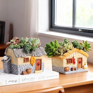 May include: Two house-shaped resin planters with textured stone-look bases and thatched roofs, each filled with green succulents. The left planter features a small white cat, while the right planter displays red chili peppers and a small white duck.