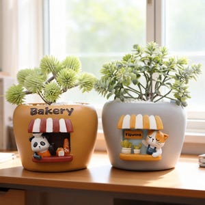 May include: Two rounded ceramic planters with artificial green plants. The left mustard-yellow planter features a panda, a red-and-white striped awning, and the word Bakery. The right gray planter features a cat, a yellow-and-white awning, and the word Flowers.