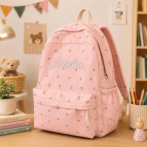 Könnte beinhalten: Ein hellrosa Rucksack mit einem wiederkehrenden Muster aus kleinen Blumen und dem in weißer Schreibschrift auf die Vordertasche gestickten Namen Amelia. Der Rucksack verfügt über einen Tragegriff, verstellbare Schultergurte und Seitentaschen.