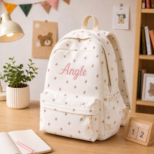 Könnte beinhalten: Ein cremefarbener Rucksack mit einem sich wiederholenden Muster aus kleinen rosa Blumen und grünen Blättern. Auf der Vorderseite ist der Name Angle in rosa Schreibschrift eingestickt. Er verfügt über einen Tragegriff, eine Reißverschlusstasche vorne und Seitentaschen.