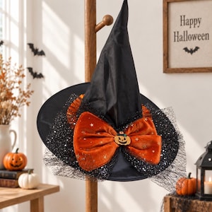 May include: A black pointed witch hat featuring a large orange velvet bow adorned with small gold star patterns and a central jack-o'-lantern emblem. The hat is decorated with layers of black glittery tulle netting around the brim.
