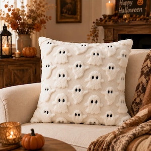 May include: A square throw pillow featuring a cream-colored knit base with a repeating pattern of raised, fuzzy white ghost figures. Each ghost has two small, dark, embroidered eyes. The texture of the ghosts contrasts with the flat knit fabric.