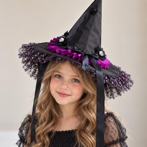 May include: A black pointed hat featuring a wide brim with purple glittery tulle trim, a pleated purple ribbon band, and small white skull accents. The hat includes long black satin ribbons hanging from the sides and a single black rose at the top.