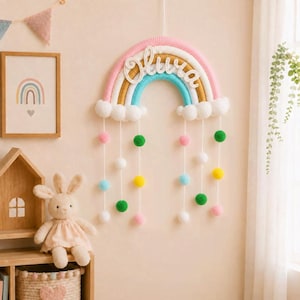 Könnte beinhalten: Eine Wanddekoration in Regenbogenform aus gewickeltem Garn in Rosa-, Beige- und Türkistönen mit weißen Pompon-Wolken an der Basis. Der Name Olivia ist in weißer Schreibschrift in der Mitte angebracht, ergänzt durch hängende bunte Pompons.
