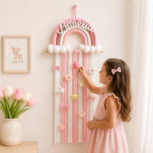May include: A wall-hanging rainbow organizer featuring the name Amelia in white script. The rainbow is crafted from pink, white, and gold cord with white pom-pom clouds. Multiple pink and white fabric strips hang below, adorned with various hair clips.