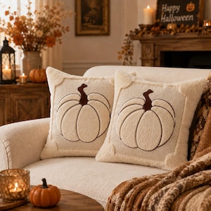 May include: Two square throw pillows with a cream-colored knit texture, each featuring a large, centered embroidered pumpkin design in dark brown thread. The pillows have a soft, plush border detail and are positioned side by side on a sofa.
