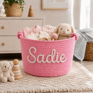 May include: A pink woven rope storage basket featuring the name Sadie in white felt lettering on the front. The basket is filled with baby clothing and a plush rabbit toy, with a small teddy bear and wooden stacking rings placed nearby.