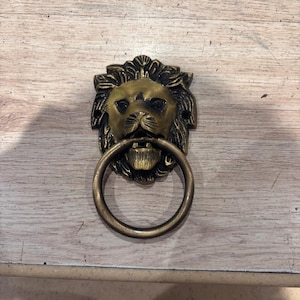 Könnte beinhalten: Ein messingfarbener Türklopfer aus Metall mit einem detaillierten Löwenkopf-Design, einer ausgeprägten Mähne und einem kreisförmigen Ring, der im Maul des Löwen gehalten wird. Das Stück hat zwei kleine Befestigungslöcher an den Seiten.