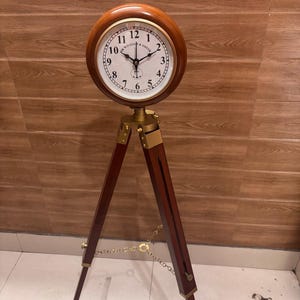 May include: A floor clock featuring a round wooden frame with a white face, black numerals, and the text Sir William & Smith. The clock is mounted on a dark brown wooden tripod stand with brass-coloured metal accents and a central chain connector.