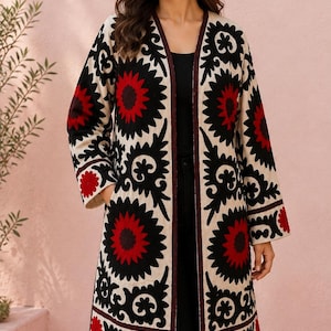 May include: A long-sleeved, open-front coat featuring a cream-coloured base with large, bold floral patterns in black and deep red. The garment has a dark maroon trim along the edges and includes side pockets, showcasing intricate embroidery throughout.