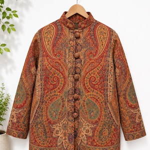 Könnte beinhalten: Eine langärmelige Jacke mit einem lebendigen, mehrfarbigen Paisley-Muster in Rot-, Orange-, Gold- und Petroltönen. Das Kleidungsstück verfügt über einen hohen Stehkragen und einen Frontverschluss mit fünf passenden, stoffbezogenen Kugelknöpfen.