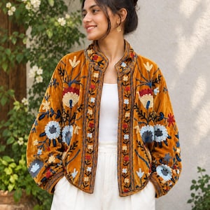 Könnte beinhalten: Eine senfgelbe Samtjacke mit offener Vorderseite und Stehkragen. Das Kleidungsstück ist mit aufwendigen Blumenstickereien in Blau-, Rot- und Cremetönen verziert, die die Ärmel und die Vorderteile der Jacke bedecken.