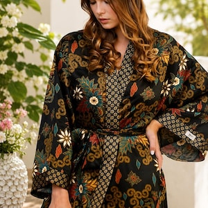 Puede incluir: Una bata tipo kimono de color negro con un estampado floral y de espirales multicolor en tonos rojo, naranja, azul verdoso y dorado. La prenda incluye un cuello ancho y puños con un estampado geométrico en contraste y un cinturón de tela.