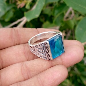 May include: A silver-toned ring featuring a large, rectangular, faceted blue gemstone set in the center. The band is crafted with intricate, embossed geometric and knotwork patterns along the sides, providing a textured, metallic finish.