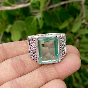 May include: A silver-toned ring featuring a large, rectangular, light green gemstone set in the center. The band of the ring is wide and decorated with intricate, raised scrollwork patterns that extend along the sides of the metal setting.