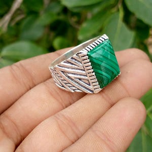 May include: A silver-toned ring featuring a rectangular, green malachite stone with distinct banded patterns. The metal band displays an intricate, textured geometric design with diagonal lines and vertical grooves surrounding the central stone.