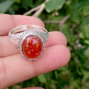 May include: A silver ring featuring a large, oval-shaped, translucent orange amber gemstone set in a textured, pitted metal band. The stone displays internal golden inclusions and a polished surface, while the surrounding metal has a dark, rugged finish.