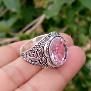 May include: A silver-toned ring featuring an oval-cut, light pink faceted gemstone set in a band with intricate, raised metalwork patterns. The band displays a textured, braided design along the edges and ornate, swirling details on the sides.