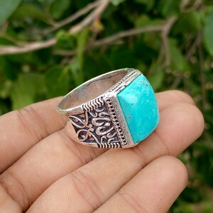 May include: A silver-toned ring featuring a large, rectangular, bright turquoise stone set in a metal band. The band displays intricate, raised, dark-toned scrollwork patterns along the sides, providing a textured contrast to the smooth stone.