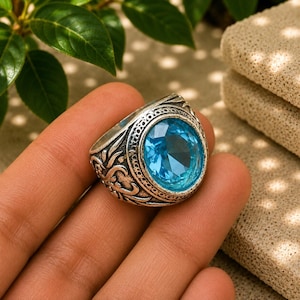Op de afbeelding: Een zilverkleurige ring met een ovaalvormige, gefacetteerde lichtblauwe edelsteen in het midden. De metalen band heeft een ingewikkeld, verhoogd krulpatroon en een getextureerde rand rondom de zetting van de centrale steen.