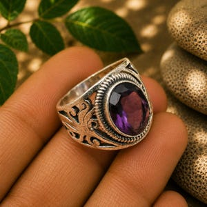 Op de afbeelding: Een zilverkleurige ring met een ovaalvormige, gefacetteerde paarse edelsteen in een decoratieve zetting. De band vertoont ingewikkelde, verhoogde krulpatronen aan de zijkanten en een gedraaide touwtextuur rondom de centrale steen.