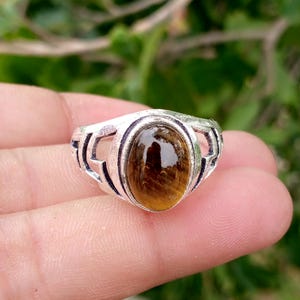 May include: A silver-toned ring featuring an oval-shaped, polished brown gemstone set in the center. The metal band displays an intricate, openwork geometric pattern that extends from the central stone setting along the sides of the ring.