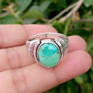 Op de afbeelding: Een zilveren ring met een grote, ovaalvormige turkoois steen met natuurlijke bruine aders. De ringband is gemaakt van gepolijst zilver en is versierd met een ingewikkeld, herhalend Grieks sleutelpatroon dat aan beide zijden is gegraveerd.