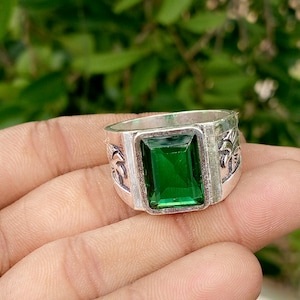 May include: A silver-toned ring featuring a large, rectangular, emerald-green faceted gemstone set in the centre. The band of the ring displays detailed engravings of palm trees on both sides of the central stone setting, creating a textured look.