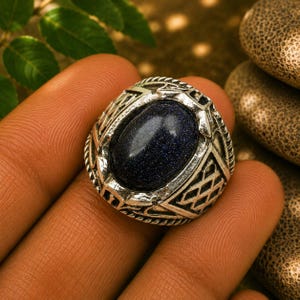 Könnte beinhalten: Ein silberfarbener Ring mit einem großen, ovalen, dunkelblauen Sandstein-Cabochon mit funkelnden Einschlüssen. Das Metallband zeigt ein kompliziertes, strukturiertes Gittermuster, das den zentralen Stein in einer breiten Fassung umgibt.