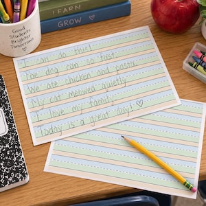 May include: Two sheets of white paper with horizontal lines in alternating light green and light blue colours, featuring dashed centre lines for handwriting practice. One sheet contains handwritten sentences including I can do this and Today is a great day.
