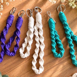 May include: Six macrame wristlet keychains arranged in pairs of purple, white, and teal. Each keychain features a twisted cord pattern, a knotted loop, and a silver-toned metal lobster clasp attached to a circular split ring for secure key attachment.