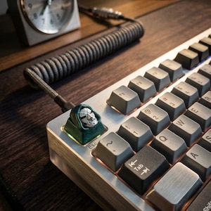 May include: A mechanical keyboard featuring a metallic silver finish and dark gray keycaps. The escape key is replaced by a custom artisan keycap shaped like a silver-colored masked figure wearing a dark green hood, connected by a coiled cable.