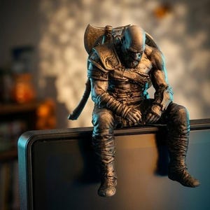 May include: A grey, metallic-toned figurine of a muscular, bald warrior with a beard, sitting with a hunched posture. The figure wears detailed armor, arm wraps, and boots, with a large axe strapped to the back, perched on the edge of a flat surface.