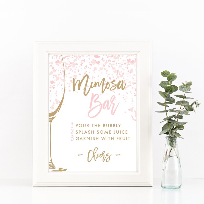 May include: A white framed sign with the words "Mimosa Bar" in gold and pink script. The sign includes instructions: "Pour the bubbly, splash some juice, garnish with fruit." A gold champagne glass outline is on the left. A small vase with greenery is on the right.