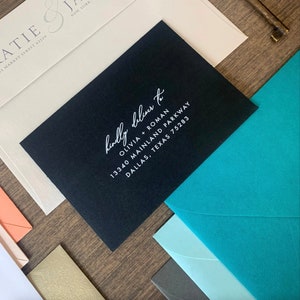 May include: A black envelope with a white handwritten address label that reads "kindly deliver to Olivia + Roman 13340 Mainland Parkway Dallas, Texas 75283".