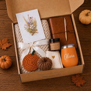 May include: A gift box containing a beige knit blanket tied with a white ribbon, a brown leather journal with a gold pen, a scented candle, a pair of white socks with a gold heart, and an orange tumbler with the name Olivia printed in white.