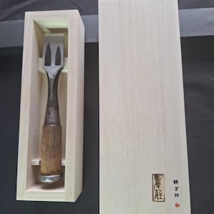 May include: A Japanese wood chisel with a dark metal blade and a wooden handle, resting inside a light-colored wooden storage box. The box lid is placed beside it, featuring a black rectangular stamp and Japanese text in black and red ink.