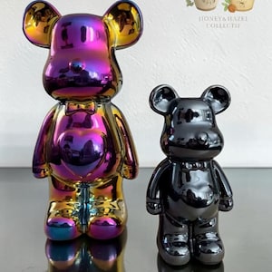 Puede incluir: Dos figuras con forma de oso y superficies lisas y reflectantes. El oso más grande tiene un acabado iridiscente multicolor con un corazón en el pecho, mientras que el más pequeño tiene un acabado negro sólido. Ambos incluyen un detalle de pajarita.