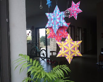 May include: A collection of colorful origami stars hanging from the ceiling. The stars are made of paper and are in various shades of pink, blue, yellow, and red. The stars are hanging in a window, with a bicycle visible in the background.