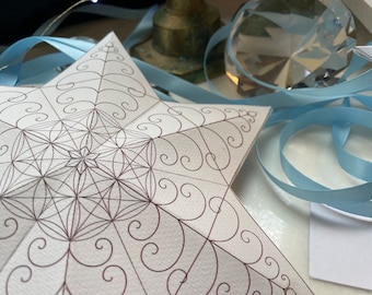 Könnte beinhalten: Ein weißer Papierstern mit einem braunen geometrischen Muster. Der Stern ist gefaltet und hat eine dreidimensionale Form. Der Stern ist von einem blauen Band umgeben.