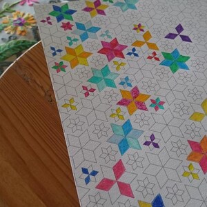 Pode incluir: Um desenho geométrico parcialmente colorido em papel. O desenho apresenta um padrão repetitivo de formas de estrelas cinzentas e flores coloridas em forma de estrela em tons de rosa, azul, amarelo, verde e roxo. O papel está sobre uma mesa de madeira.